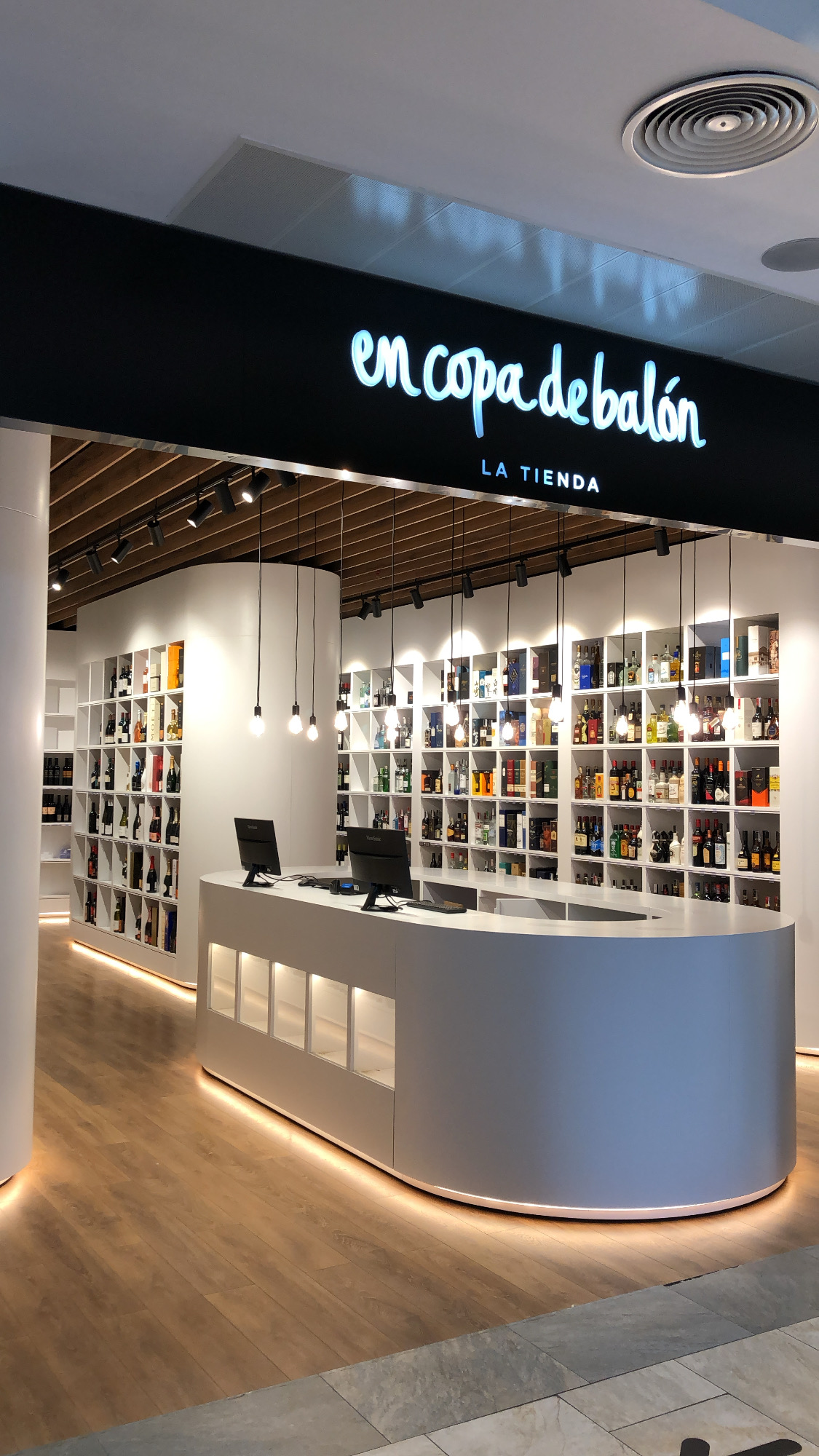 General view of the shop with a white oval counter, wine and spirits shelving and hanging bulbs — En Copa de Balón