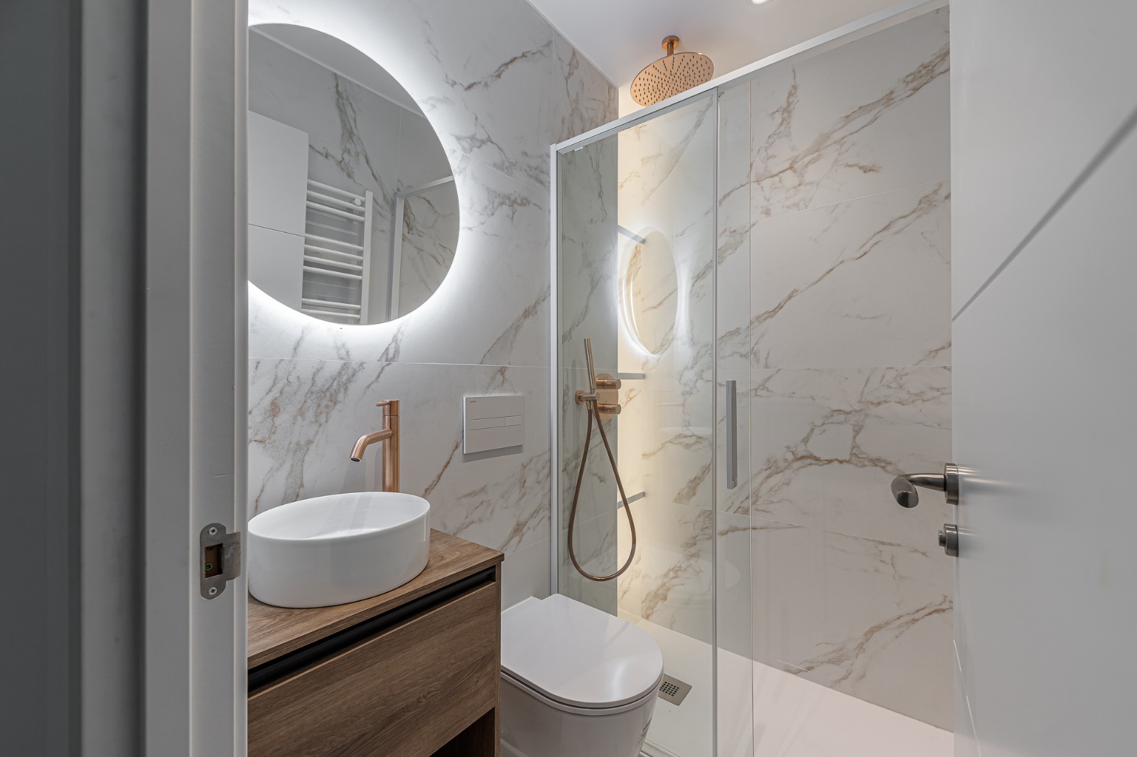 White marble bathroom with round backlit mirror and copper fixtures