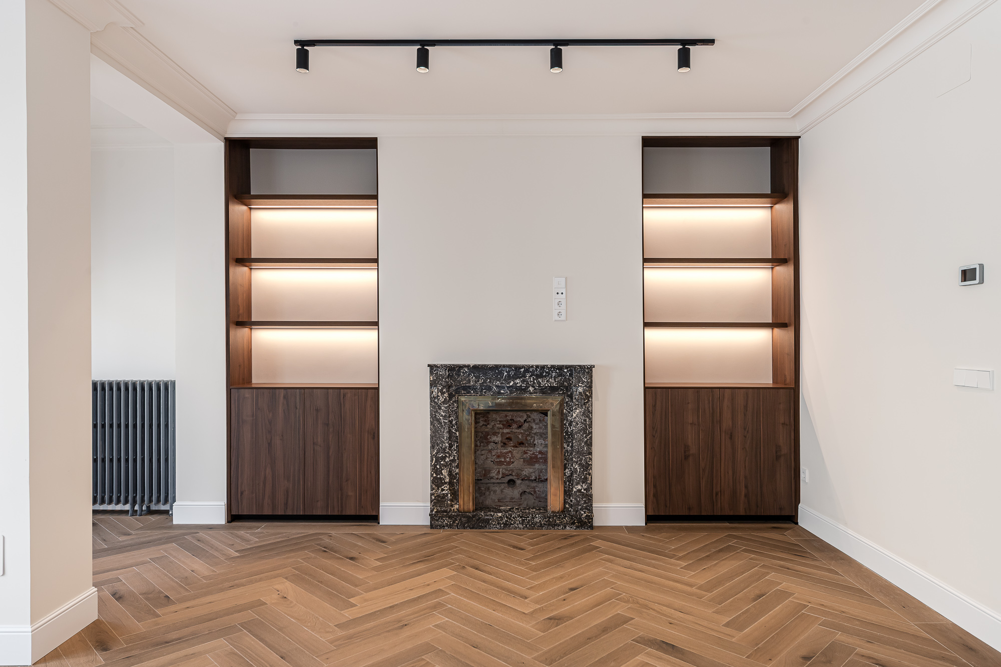 Living room with herringbone wood flooring, built-in wood shelving with integrated lighting and a fireplace with a dark stone surround — Lucio del Valle Residence
