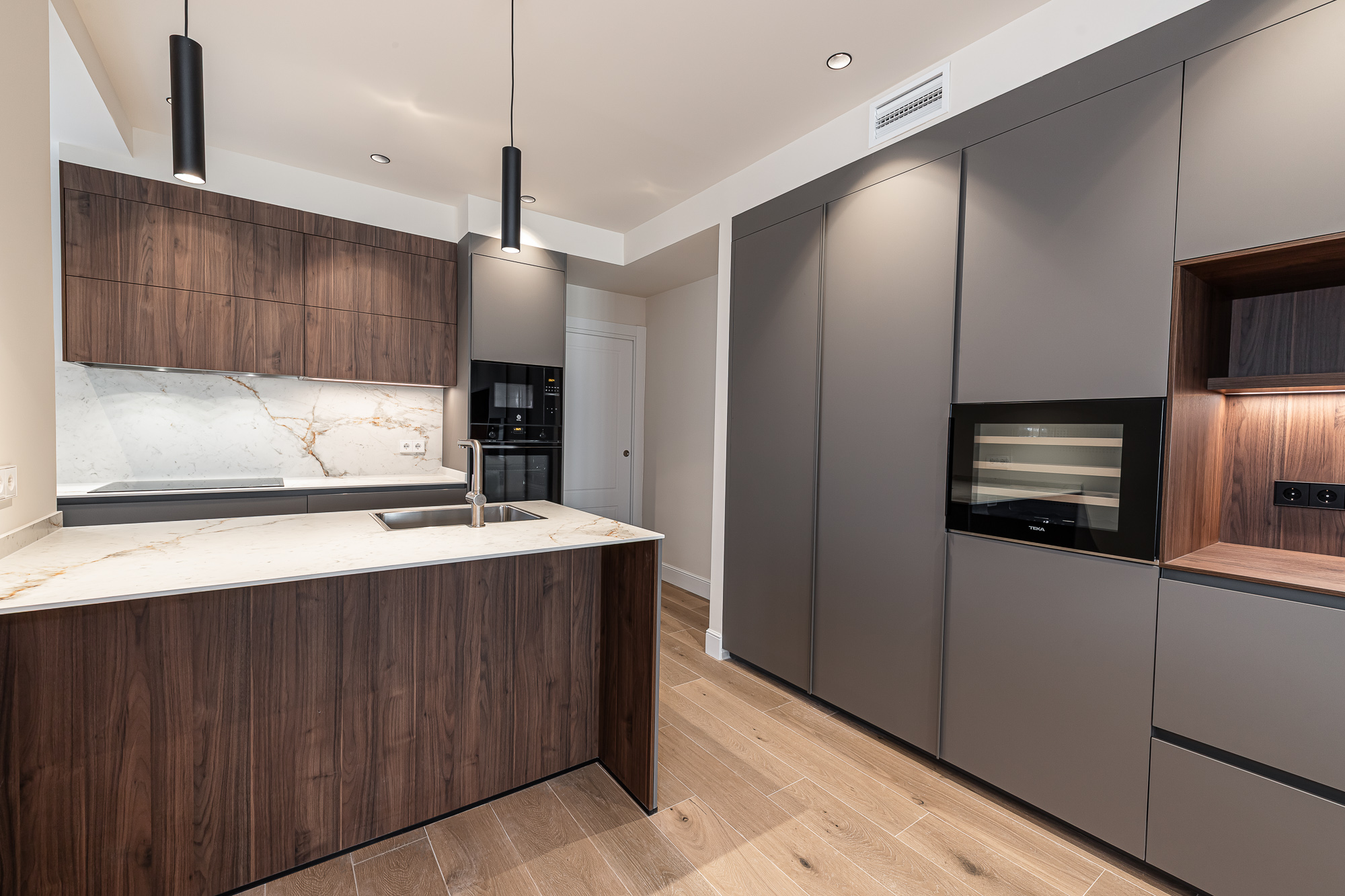 Kitchen with a dark wood island, light veined backsplash and tall grey cabinets with integrated appliances