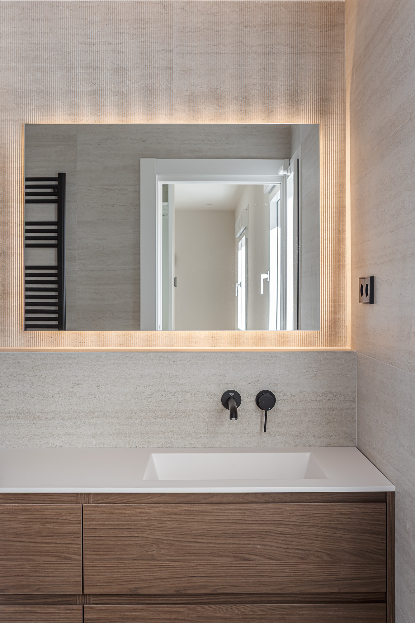 Wood bathroom vanity with white countertop, black wall-mounted taps and backlit mirror