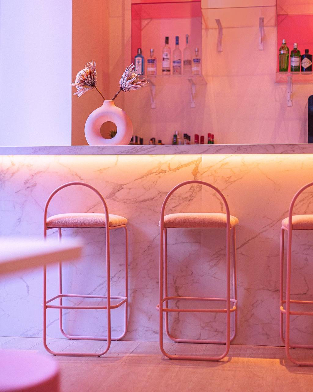 Bar with a white veined front, pink stools and bottle shelves with indirect lighting