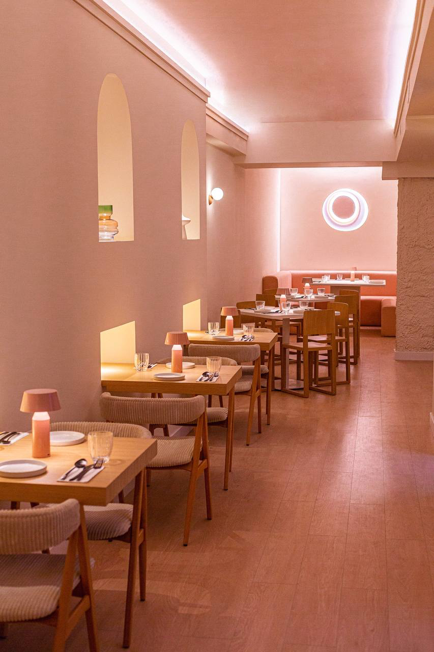 Long dining room with arched wall niches, wooden tables and an orange banquette at the back beneath a circular light fitting