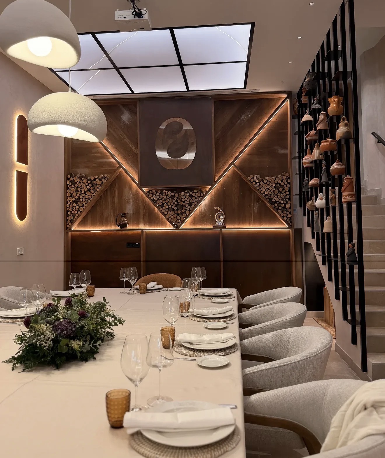 Dining room with a long dark wood table laid for a meal, upholstered armchairs in light fabric, white pendant lamps and a wood wall with linear lighting — Olar by O'Grelo Restaurant
