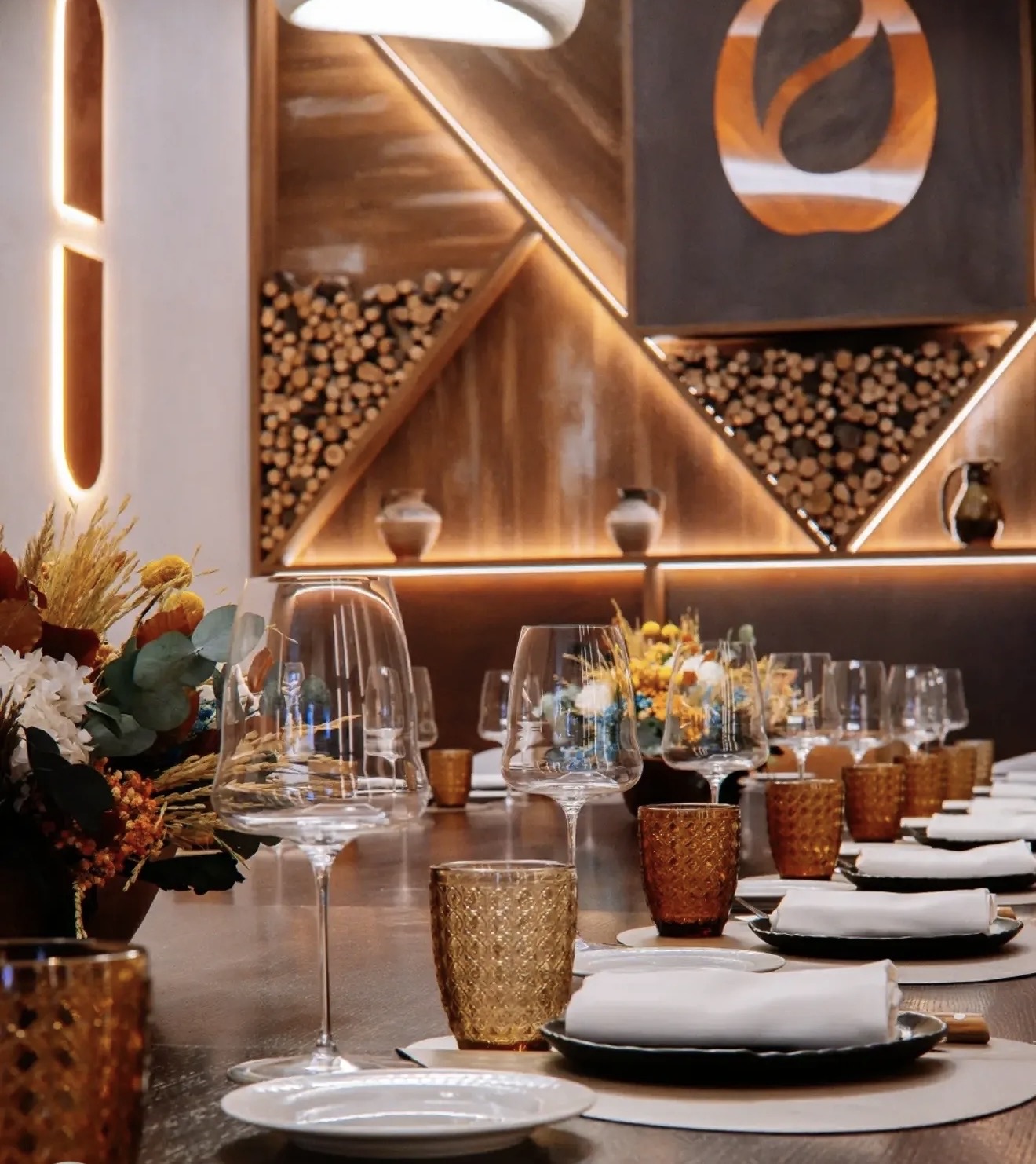 Dining room with a long laid table, a wood wall with niches of stacked firewood and, on the right, a staircase with ceramic pieces on a black metal frame