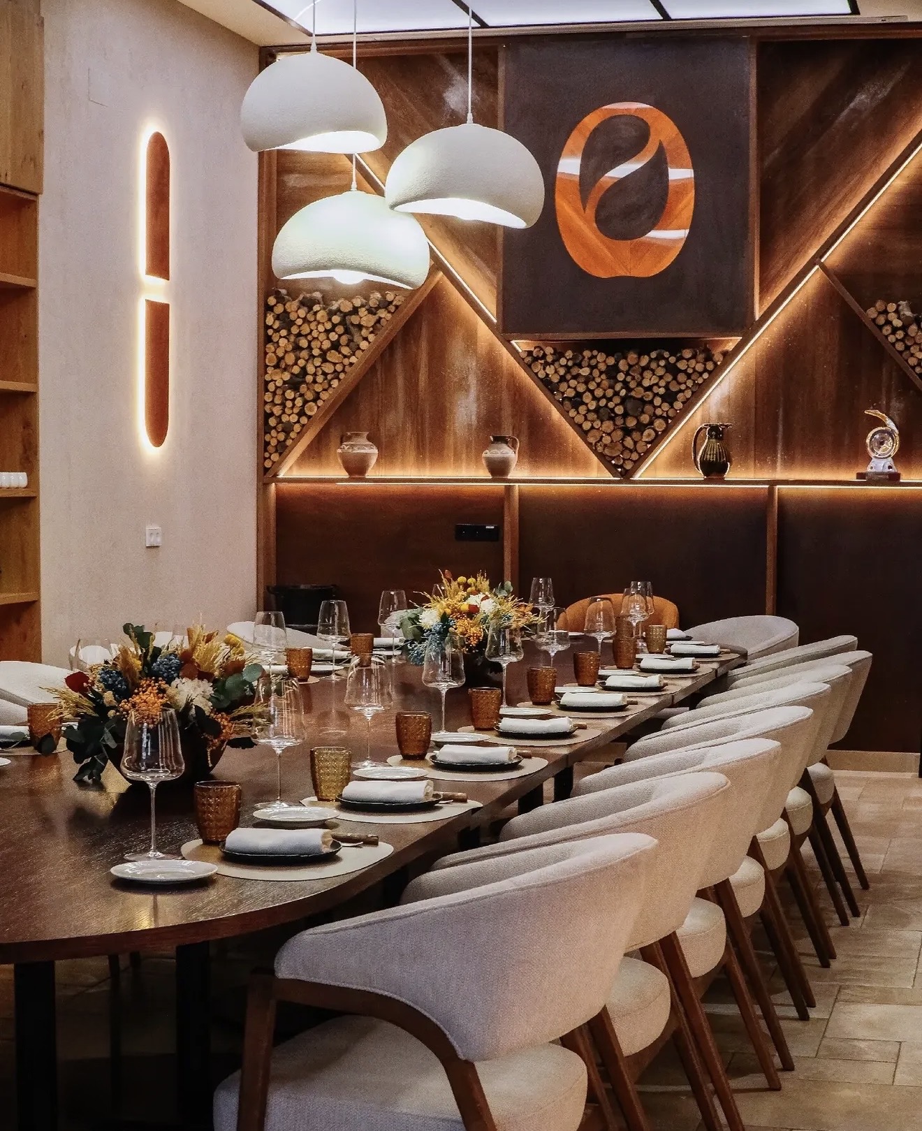 View along the table with white pendant lamps, upholstered armchairs and a wood wall with indirect lighting