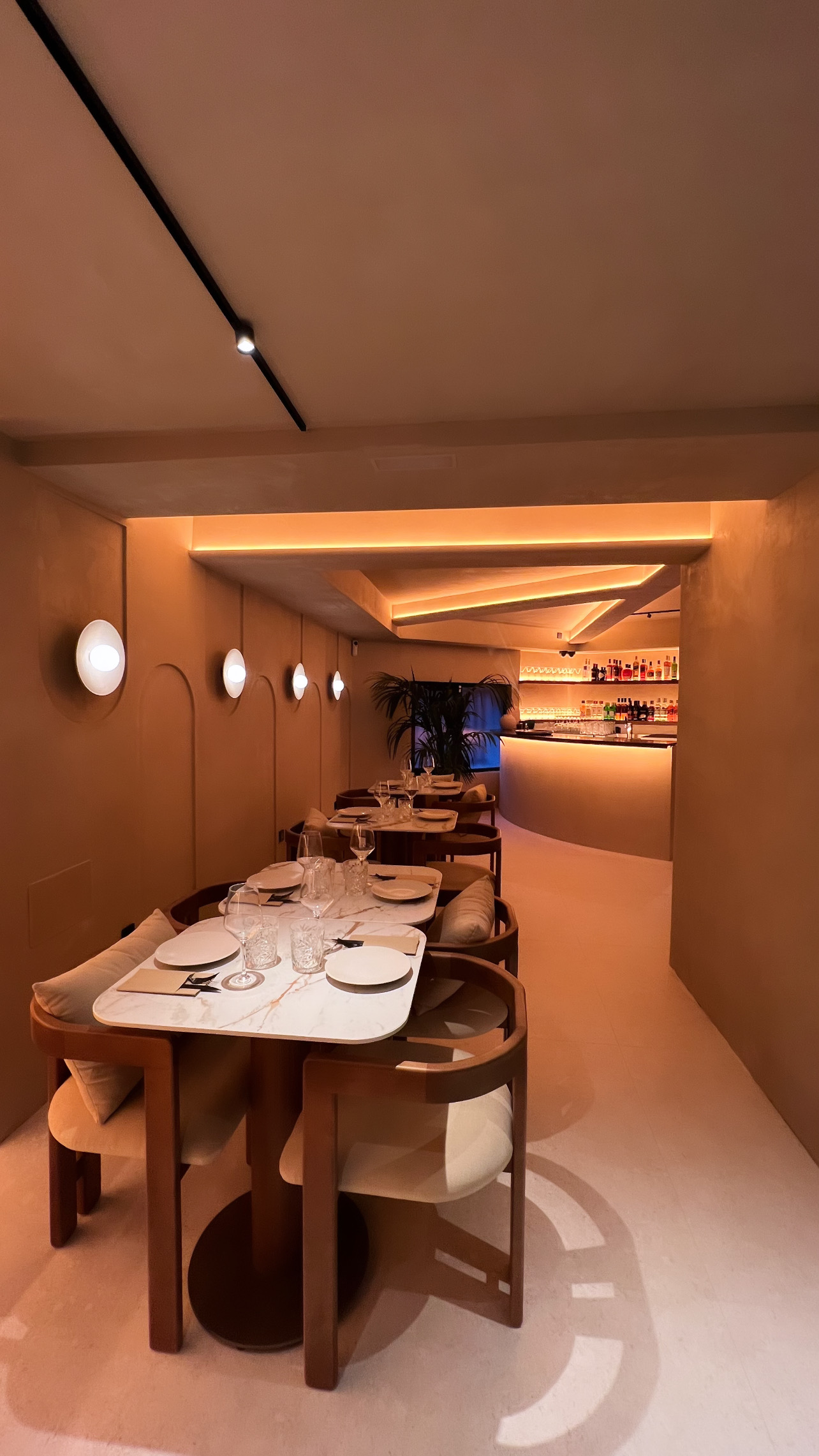 Narrow dining room with laid tables, circular wall lights on a wall with raised arches and the bar at the back