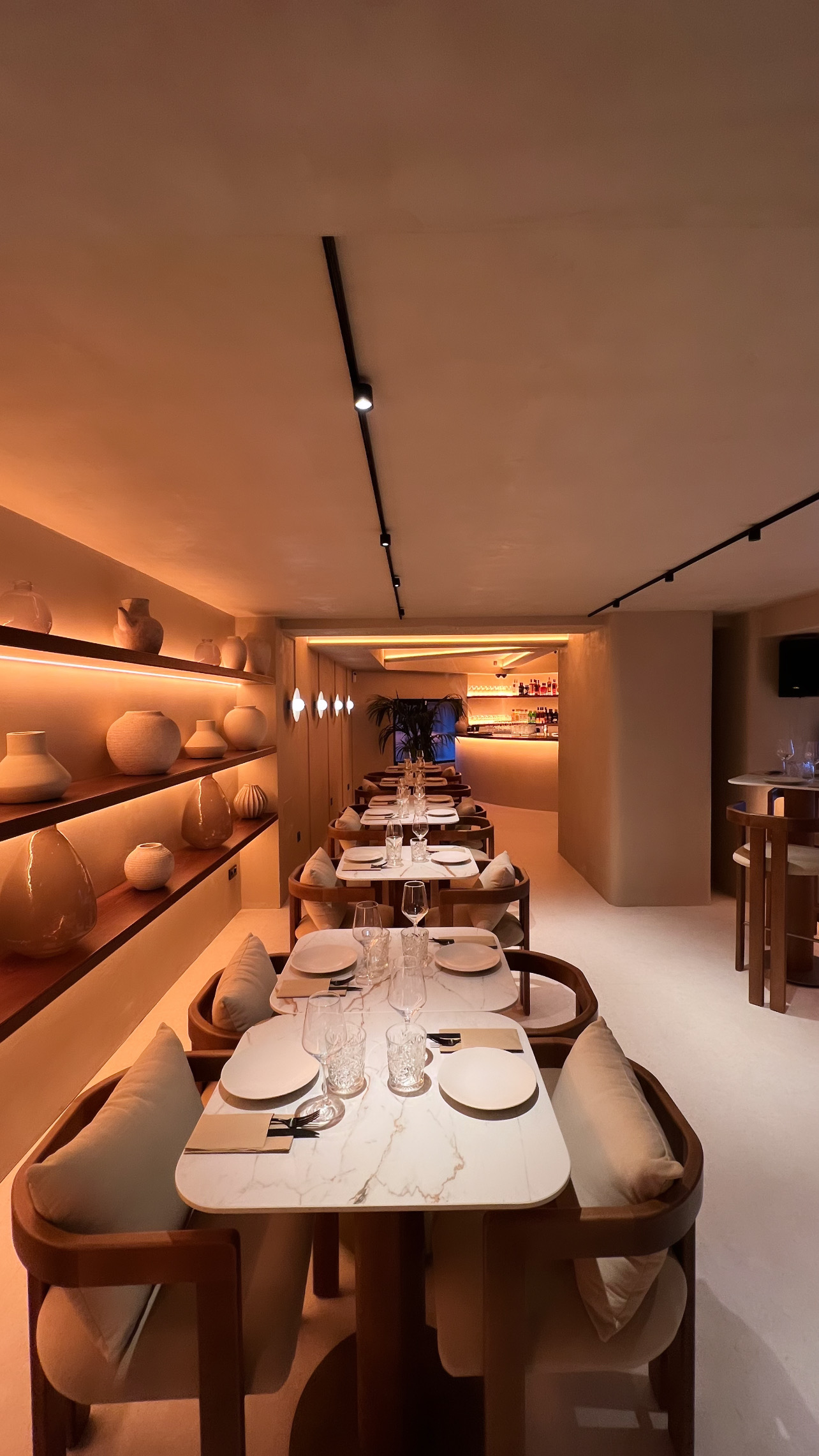 Row of tables with white veined tops and wooden armchairs, with a lit ceramics shelf on the left and the bar at the back — Santiago Ultramarinos Restaurant